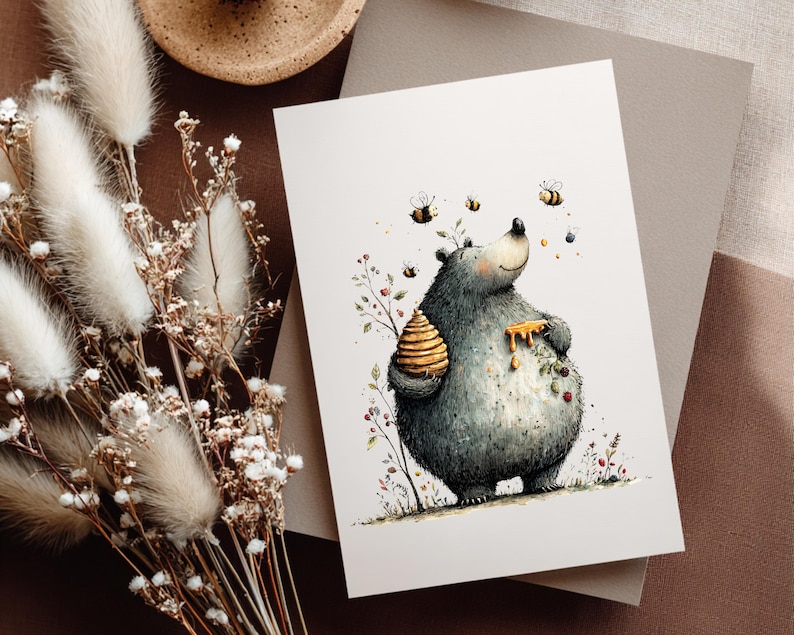 May include: A watercolour illustration of a bear with a beehive and honey dripping down its chest. Bees fly around the bear, and the artwork is on a white card. The card is on a brown surface with dried flowers.