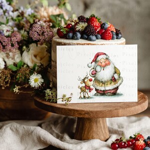 K&ouml;nnte beinhalten: Eine festliche Szene mit einem Kuchen, der mit Beeren belegt ist, einem Blumenarrangement und einer Gru&szlig;karte. Die Karte zeigt den Weihnachtsmann und ein Rentier. Der Kuchen steht auf einem Holzst&auml;nder, mit Beeren im Vordergrund.