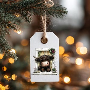 May include: A white wooden tag with a watercolor illustration of a Highland cow peering out of a window, adorned with a small wreath. The tag is suspended by a natural twine string and a brass eyelet, perfect for holiday decor.