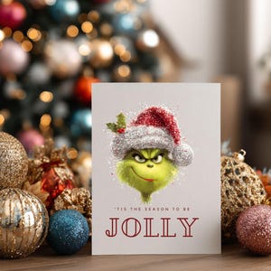 Peut inclure: Carte de No&euml;l avec un personnage de dessin anim&eacute; vert portant un bonnet de P&egrave;re No&euml;l. Le texte "'Tis the season to be JOLLY" est en rouge. Le fond pr&eacute;sente des boules de No&euml;l et un sapin flou.