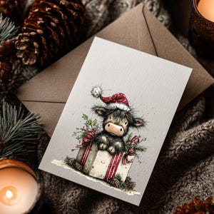 May include: A Christmas greeting card featuring a Highland cow wearing a Santa hat, sitting in a gift box with a red ribbon. The card is on a neutral background with pine cones, greenery, and a lit candle.