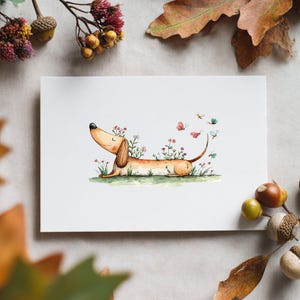 May include: A watercolour illustration on a white card depicts a dachshund dog lying in grass, adorned with flowers and butterflies. The dog is light brown with a long body and floppy ears. The background includes autumn leaves and acorns.