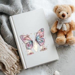 May include: A white journal with a butterfly design. The butterfly wings are pastel colors with floral accents. A small teddy bear with a gray scarf sits next to the journal. A gray knitted scarf is in the background.