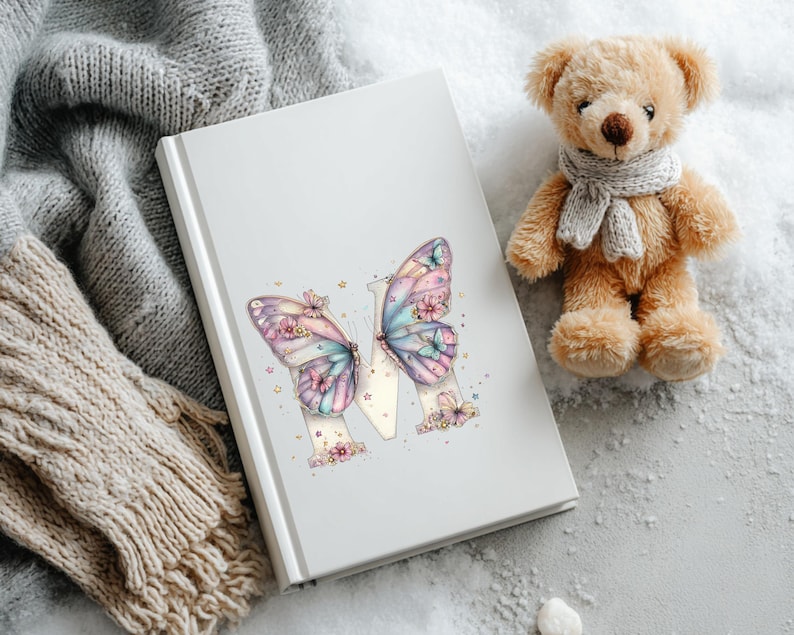 May include: A white journal with a butterfly design. The butterfly wings are pastel colors with floral accents. A small teddy bear with a gray scarf sits next to the journal. A gray knitted scarf is in the background.