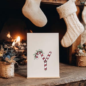 May include: A festive print showing the letter 'Y' styled as a candy cane with red and white stripes, decorated with green leaves and berries. The print is on a wooden stand, with a fireplace and Christmas stockings in the background.