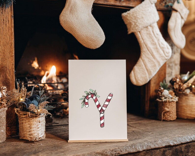 May include: A festive print showing the letter 'Y' styled as a candy cane with red and white stripes, decorated with green leaves and berries. The print is on a wooden stand, with a fireplace and Christmas stockings in the background.