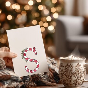 May include: A greeting card with a candy cane-style letter "S" in red and white stripes, adorned with a green leaf. A steaming mug of a drink is next to the card. The background is blurred with Christmas lights.