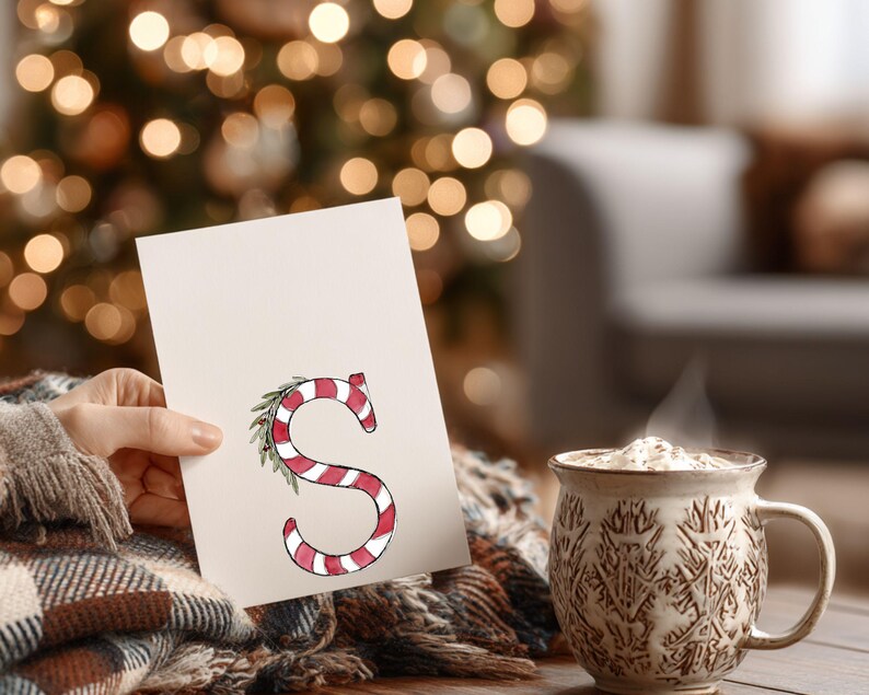 May include: A greeting card with a candy cane-style letter "S" in red and white stripes, adorned with a green leaf. A steaming mug of a drink is next to the card. The background is blurred with Christmas lights.