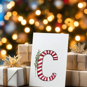 May include: A festive Christmas image showing a white card with a red and white striped letter 'C' design, adorned with green leaves. The card is in front of wrapped gifts with gold ribbon and bows, set against a blurred backdrop of Christmas lights.