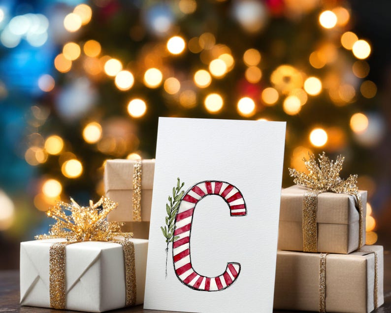 May include: A festive Christmas image showing a white card with a red and white striped letter 'C' design, adorned with green leaves. The card is in front of wrapped gifts with gold ribbon and bows, set against a blurred backdrop of Christmas lights.