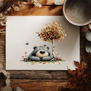 May include: A watercolour illustration of a bear resting under a tree with autumn leaves. The bear is grey with a tan snout and is surrounded by mushrooms, bees, and fallen leaves. The artwork is on white paper, set on a wooden surface.