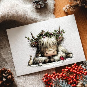 May include: A watercolor illustration of a Highland cow wearing a holly wreath, surrounded by small birds and a rabbit. The cow is lying in snow, with a red bird perched nearby. A festive holiday card.