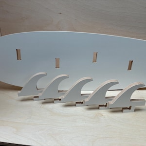 Könnte beinhalten: Ein Surfbrett-Rack aus Holz in Hellgrau. Es hat ein ovales Brett mit Schlitzen und fünf wellenförmigen Finnenhaltern. Das Rack ist für die Aufnahme eines Surfbretts konzipiert. Maße in Zentimetern.
