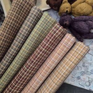 May include: A selection of rolled fabric samples in various colours and plaid patterns. The fabrics include shades of brown, green, and red, with a tan and cream-coloured fabric in the foreground. The image also shows several small teddy bears.