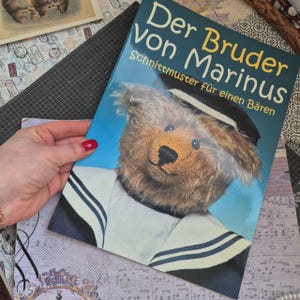 Op de afbeelding: Een softcover boek getiteld Der Bruder von Marinus Schnittmuster für einen Bären met een foto van een bruine teddybeer met een donkerblauwe matrozenmuts en een witte matrozenkraag met donkerblauwe strepen tegen een lichtblauwe achtergrond.