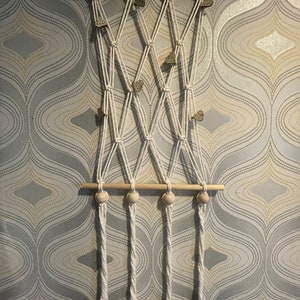 Peut inclure: Une suspension murale en macram&eacute; avec une baguette en bois et trois perles en bois. Le macram&eacute; est r&eacute;alis&eacute; avec un cordon blanc et pr&eacute;sente un motif en losange. La suspension murale est d&eacute;cor&eacute;e de petits papillons en bois.