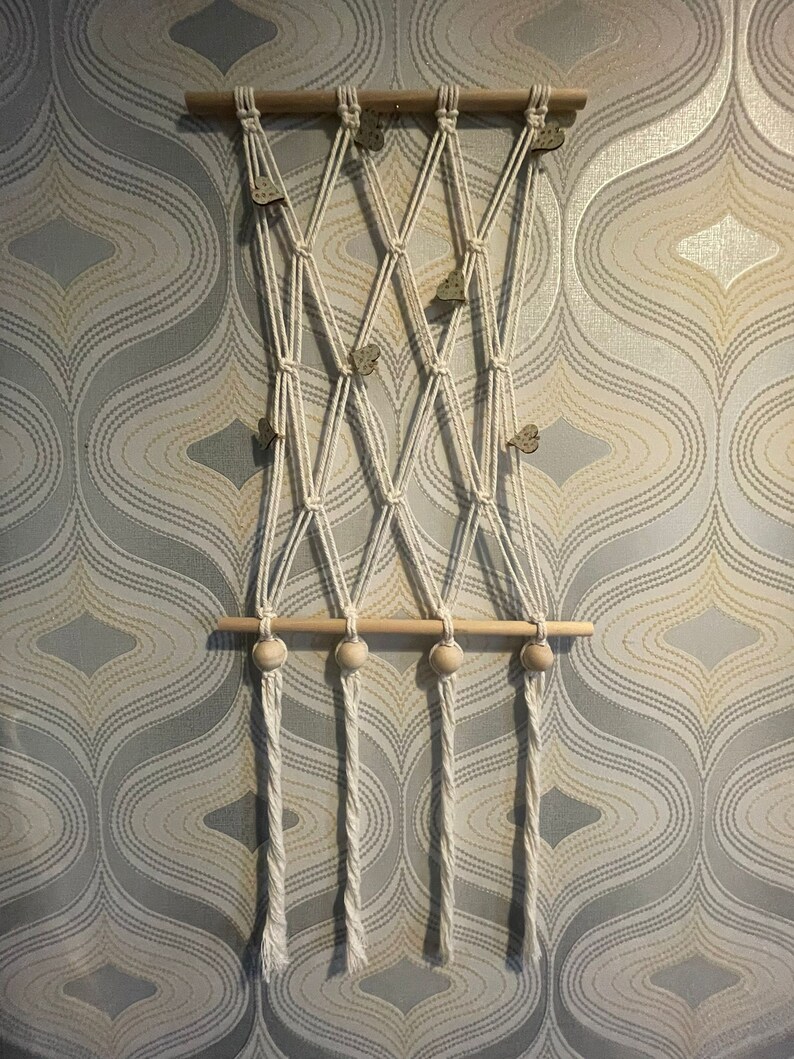 Peut inclure: Une suspension murale en macram&eacute; avec une baguette en bois et trois perles en bois. Le macram&eacute; est r&eacute;alis&eacute; avec un cordon blanc et pr&eacute;sente un motif en losange. La suspension murale est d&eacute;cor&eacute;e de petits papillons en bois.