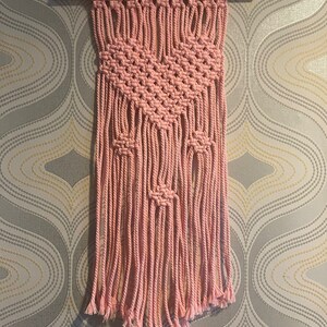 Peut inclure: Une suspension murale en macram&eacute; rose avec un motif g&eacute;om&eacute;trique et des franges. La suspension est r&eacute;alis&eacute;e en cordon de coton et est fix&eacute;e &agrave; une baguette en bois.