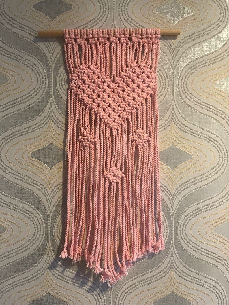 Peut inclure: Une suspension murale en macram&eacute; rose avec un motif g&eacute;om&eacute;trique et des franges. La suspension est r&eacute;alis&eacute;e en cordon de coton et est fix&eacute;e &agrave; une baguette en bois.