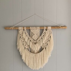 Peut inclure: Une suspension murale en macram&eacute; beige avec une tringle en bois en haut. La suspension pr&eacute;sente un motif g&eacute;om&eacute;trique avec des d&eacute;tails en franges.