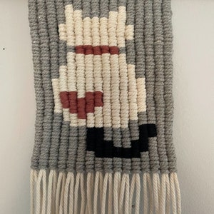 Peut inclure: Une tapisserie murale en macram&eacute; de couleur grise, blanche, rouge et noire avec un motif g&eacute;om&eacute;trique. La tapisserie est dot&eacute;e d'une baguette en bois en haut et d'une frange en bas.