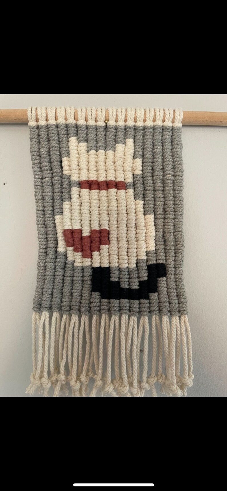 Peut inclure: Une tapisserie murale en macram&eacute; de couleur grise, blanche, rouge et noire avec un motif g&eacute;om&eacute;trique. La tapisserie est dot&eacute;e d'une baguette en bois en haut et d'une frange en bas.