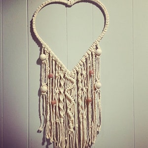 Peut inclure: Une suspension murale en macram&eacute; en forme de c&oelig;ur blanche avec des perles en bois. Le macram&eacute; est r&eacute;alis&eacute; avec un cordon blanc &eacute;pais et pr&eacute;sente une frange en bas.