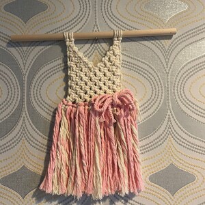 Peut inclure: Une suspension murale en macram&eacute; rose et blanche en forme de robe avec un n&oelig;ud &agrave; la taille.