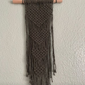 Peut inclure: Une suspension murale en macram&eacute; vert fonc&eacute; avec un motif g&eacute;om&eacute;trique et des franges. La suspension est fix&eacute;e &agrave; une baguette en bois.