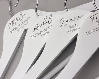 May include: Four white wooden hangers with black text. The hangers are labeled "Natalie Maid of Honour 7th June 2025", "Rachel Mother of the 7th June", "Laura Bride 7th June 20", and "T Mother of the Bride".