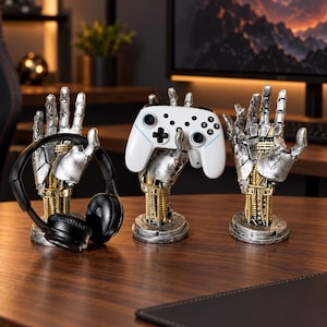 Puede incluir: Tres soportes con forma de mano robótica con acabado plateado y dorado envejecido. Un soporte sostiene auriculares negros, otro un mando de juego inalámbrico blanco y el tercero está vacío. Cada mano está montada sobre una base circular.
