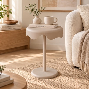Puede incluir: Una mesa auxiliar de color beige con un tablero festoneado en forma de nube y una base cilíndrica central. La mesa tiene un jarrón de cerámica beige con ramas verdes y una taza de cerámica beige sobre el libro titulado The KINFOLK Home.