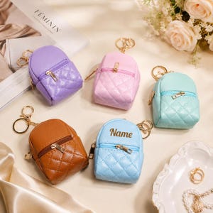 May include: Five miniature quilted backpack keychains in purple, light pink, mint green, brown, and light blue. Each features a gold-tone zipper, a metal keyring, and a front pocket. The light blue backpack has the word Name printed in dark text.