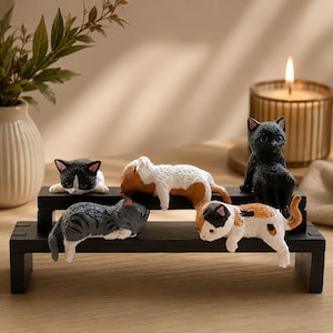 May include: A set of five small cat figurines displayed on a two-tiered black wooden stand. The cats feature various coat patterns including solid black, grey tabby, white and orange, and black and white, all in different resting or sitting poses.