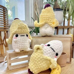 Puede incluir: Tres peluches de plátano de crochet. Dos están de pie, uno está acostado con un plátano real a su lado. Los peluches de plátano son blancos con cáscaras de plátano de crochet amarillo. Las cáscaras de plátano tienen un tallo de crochet morado. Los peluches tienen ojos negros y una boca rosa. El plátano real es amarillo y marrón.