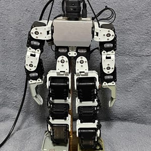 May include: A robotic figure with a silver body, black and white articulated limbs, and a black head. The robot features a complex mechanical design with visible wiring and the text "AX-12+" on its joints. It stands on a gold-coloured base.