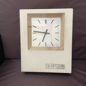 Puede incluir: Un reloj de cuarzo color crema con una esfera cuadrada y manecillas y números negros. El reloj está encerrado en un marco rectangular y tiene el texto "quartz-m" y "PATEK PHILIPPE" impreso en la parte delantera.