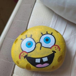 May include: A yellow painted rock featuring the face of a cartoon character with large blue eyes, a wide black mouth, two white front teeth, a pink tongue, and red cheek markings. The surface of the rock is textured with darker yellow spots.