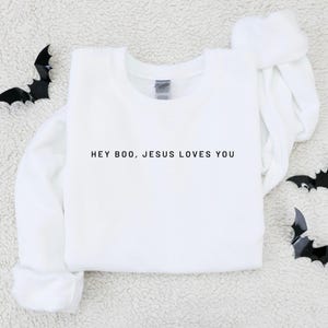 May include: White crew neck sweatshirt with the text "HEY BOO, JESUS LOVES YOU" in black. The sweatshirt is surrounded by black bat decorations, creating a Halloween theme. The fabric appears soft and comfortable.