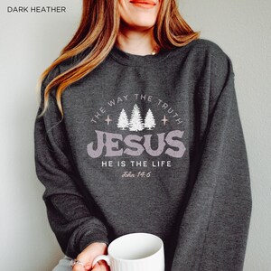 May include: Dark heather gray sweatshirt with the text "THE WAY THE TRUTH JESUS HE IS THE LIFE John 14:6" in a distressed, light purple font. The design includes three white pine trees and stars. The sweatshirt is a casual, comfortable style.
