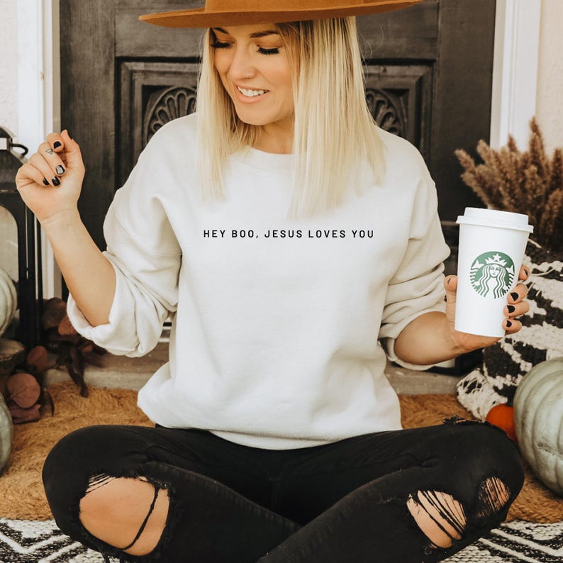 May include: A cream-colored sweatshirt with the text "HEY BOO, JESUS LOVES YOU" in black. The person is wearing a brown hat and black ripped jeans, holding a white coffee cup with a green logo.