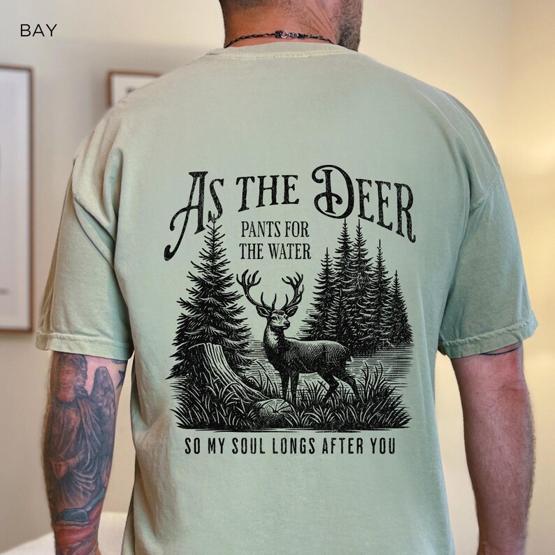 May include: A light green t-shirt with a black graphic print of a deer in a forest. The text on the shirt reads "As the Deer, Pants for the Water, So my Soul Longs After You."