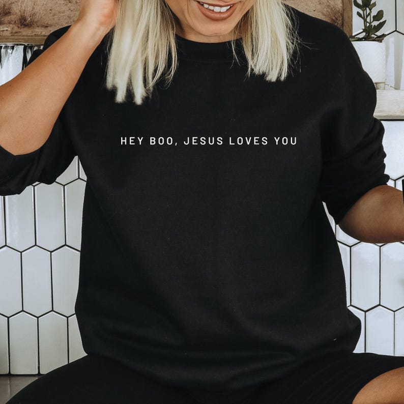 May include: Black crew neck sweatshirt with the white text "HEY BOO, JESUS LOVES YOU". The sweatshirt is a solid black color and has long sleeves. The text is centered on the front of the shirt.