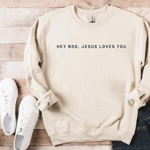 May include: A cream-colored sweatshirt with the text "HEY BOO, JESUS LOVES YOU" in black. The sweatshirt is paired with white sneakers and a pair of blue jeans. The sweatshirt has a crew neck and long sleeves.