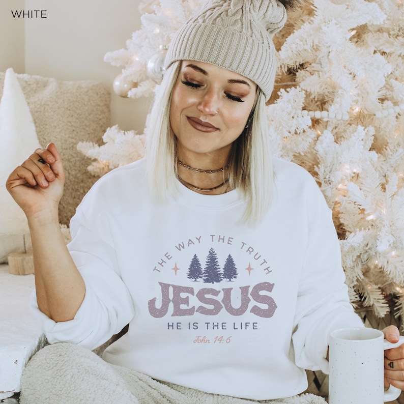 May include: White sweatshirt with the text "THE WAY THE TRUTH JESUS HE IS THE LIFE John 14:6" in a circular design. The design includes purple text and a graphic of three pine trees. The sweatshirt is a casual style.