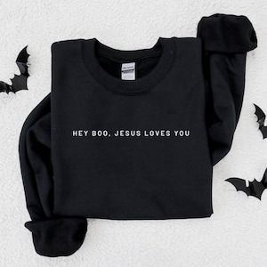 May include: Black crewneck sweatshirt with the text "HEY BOO, JESUS LOVES YOU" in white. The sweatshirt is displayed on a white, textured surface with black bat cutouts scattered around the edges.