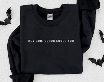 Peut inclure: Sweat-shirt noir à col rond avec l'inscription blanche "HEY BOO, JESUS LOVES YOU". Le sweat-shirt est posé sur une surface blanche texturée, avec des décorations de chauve-souris noires sur les bords.