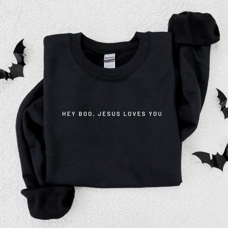 May include: Black crewneck sweatshirt with the text "HEY BOO, JESUS LOVES YOU" in white. The sweatshirt is displayed on a white, textured surface with black bat cutouts scattered around the edges.