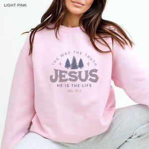 May include: Light pink sweatshirt with the text "THE WAY THE TRUTH JESUS HE IS THE LIFE John 14:6" in a gray, distressed font. The design includes three stylized pine trees. The sweatshirt is a soft, casual style.