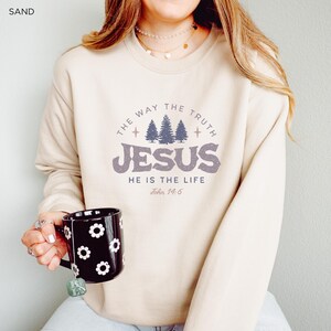 May include: Sand-colored sweatshirt with a graphic design featuring the text "THE WAY THE TRUTH JESUS HE IS THE LIFE John 14:6". The sweatshirt is paired with a black mug decorated with white flower shapes.